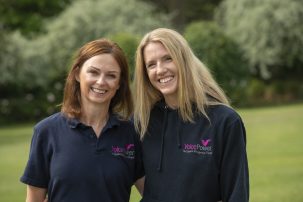 Two women stand together smiling at the camera. On the left is a shorter woman with brown hair, on the right is a woman with blonde hair. They are both wearing a VoicePower branded top and standing in a park.