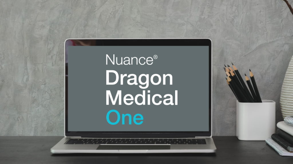 Laptop sits on desk with pens and a vase. The laptop shows a grey background with the 'Nuance Dragon Medical One' logo.
